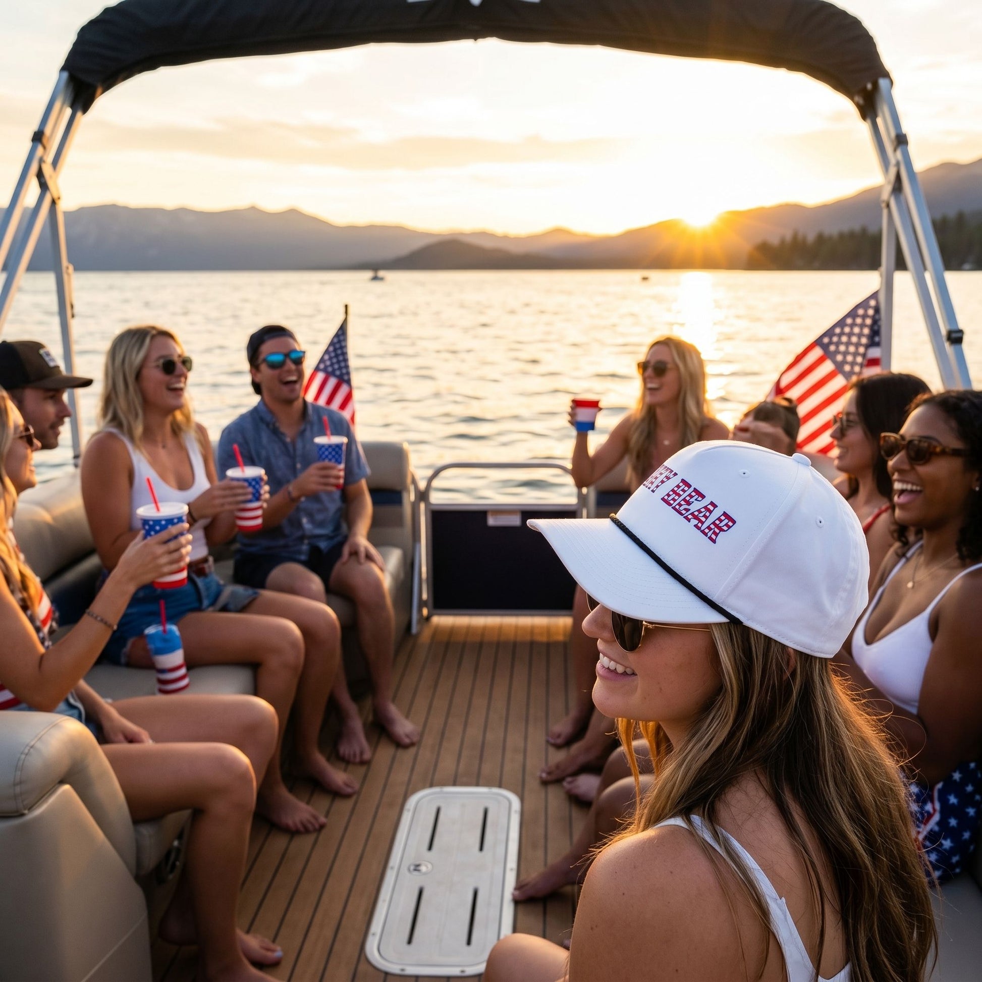 Small All-American Rope Cap on boat deck with Lake Tahoe sunset, showing embroidered flag and relaxed outdoor celebration scene.