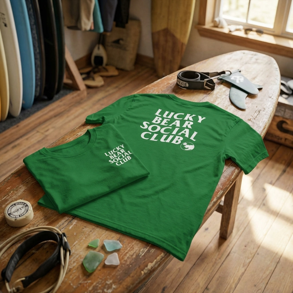Flat-lay of Lucky Bear Social Club T-shirt on surfboard in surf shop, showcasing fabric softness and logo design.