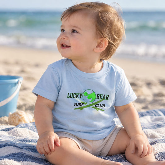 Toddler wearing a light blue cotton t-shirt sitting on a beach towel in soft natural lighting.