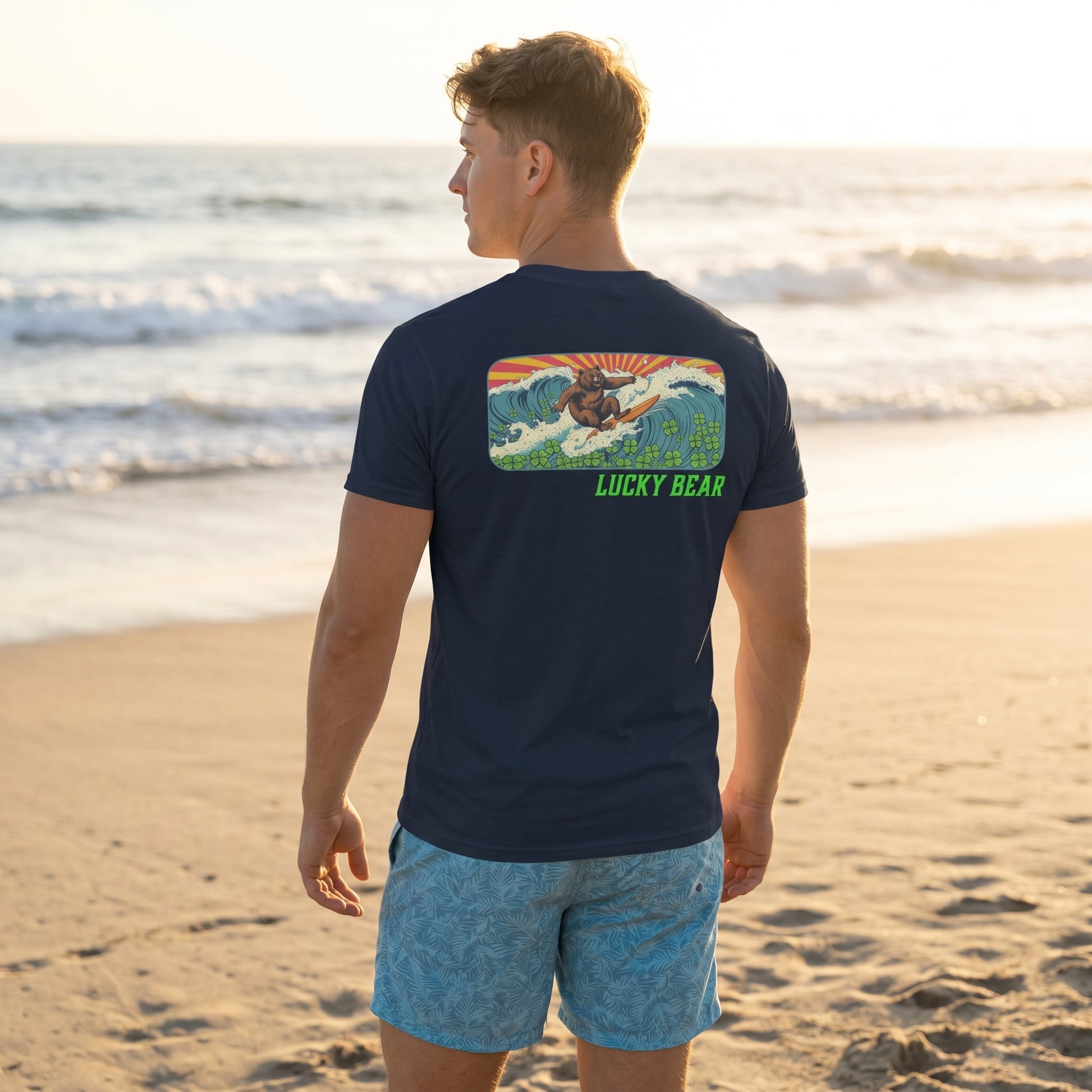 Relaxed model wearing Retro Surfing Bear Tee outdoors on sand near the ocean at golden hour.