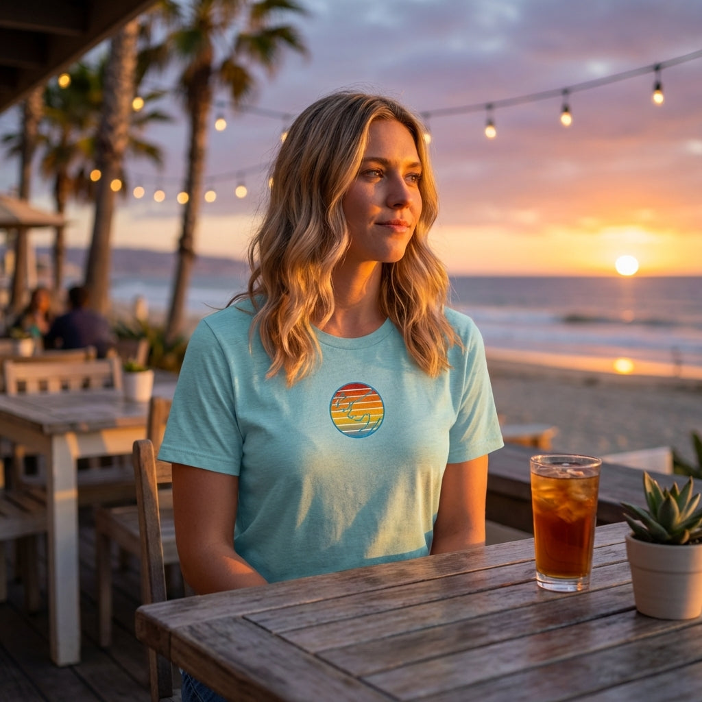 Lifestyle scene of woman at beachside cafe during sunset in relaxed fit sunset T-Shirt, highlighting vibrant colors and fabric details.