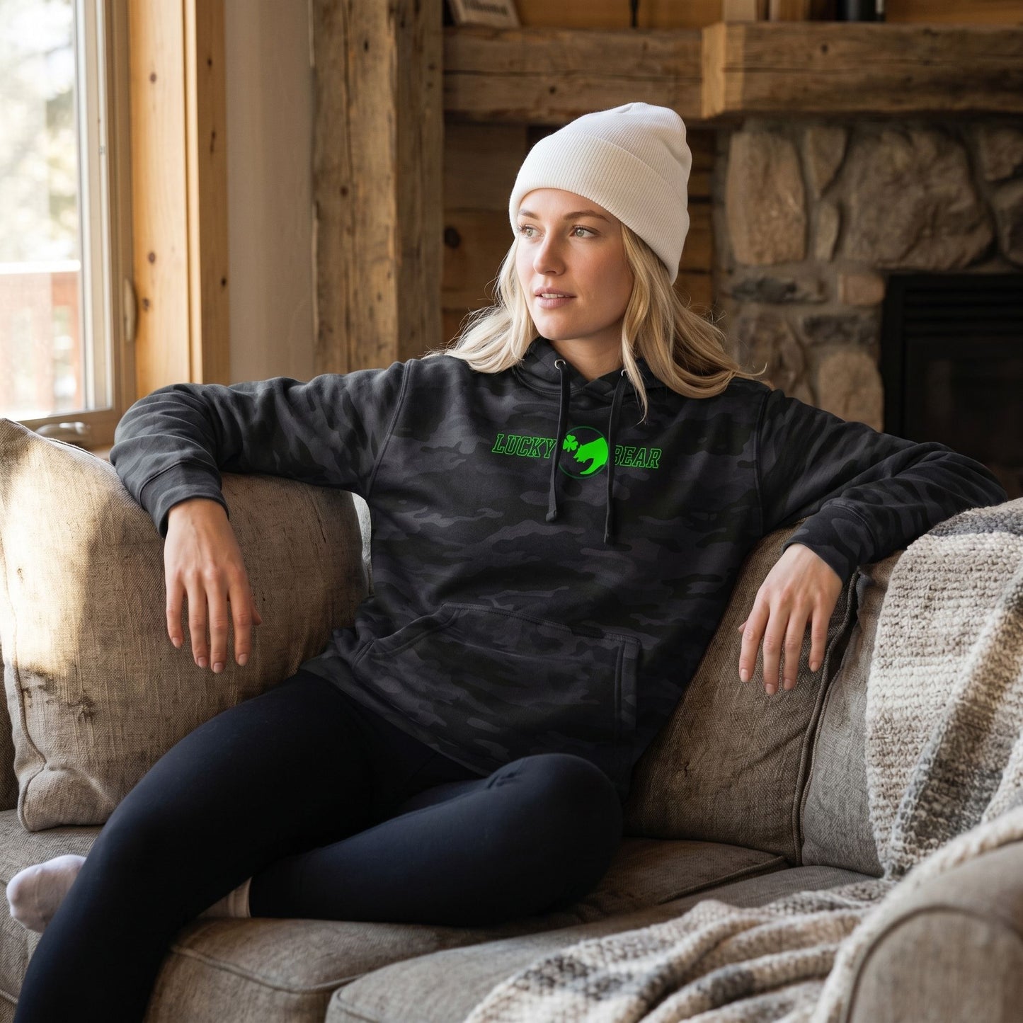 Blonde female model in a mountain cabin wearing Lucky Bear heavyweight hoodie, sitting on a cozy couch with leggings and a white beanie, highlighting its sturdy design and relaxed style.