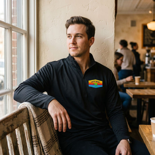 Male model in a coffee shop wearing a lightweight sunset quarter-zip pullover with highlighted zipper and collar, sitting on a rustic wooden bench.