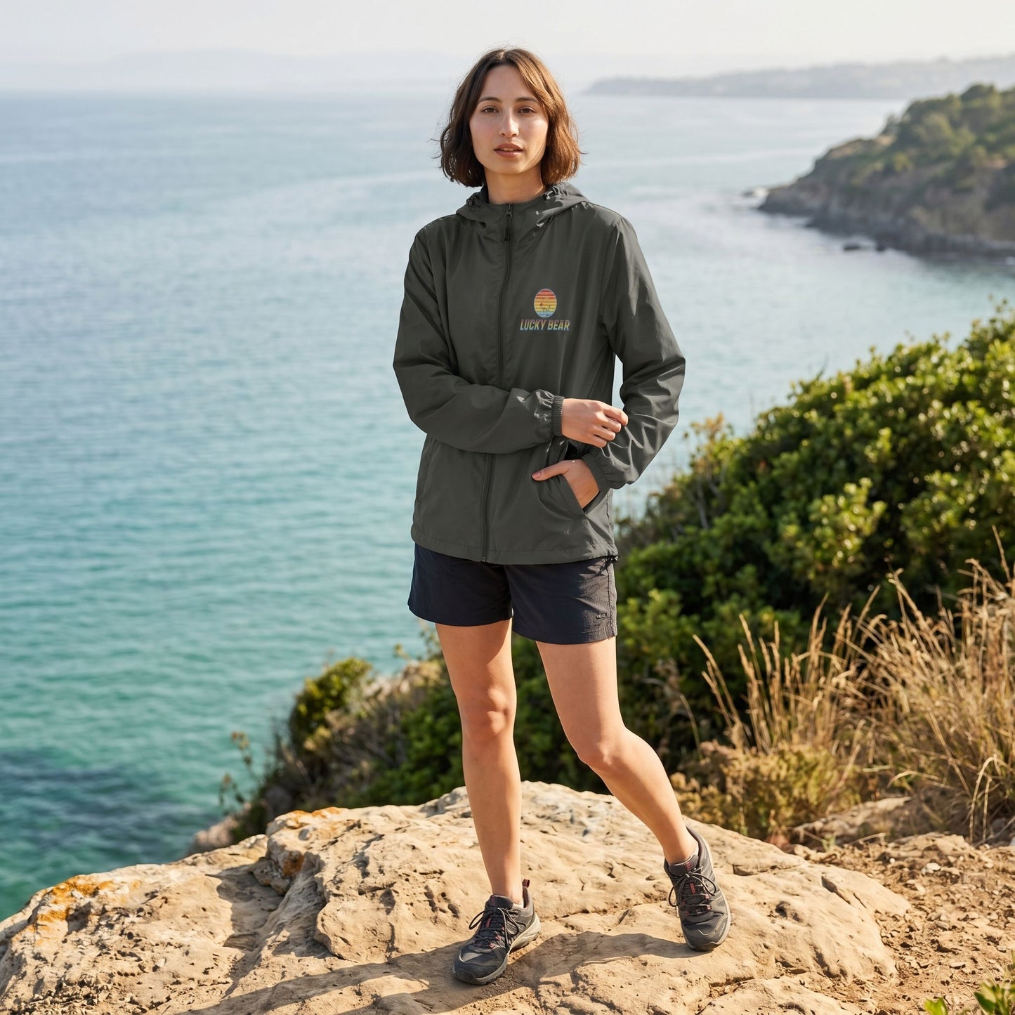 Full-body shot of a brunette female model outdoors wearing a lightweight, water-resistant windbreaker overlooking the ocean, highlighting its design and features.