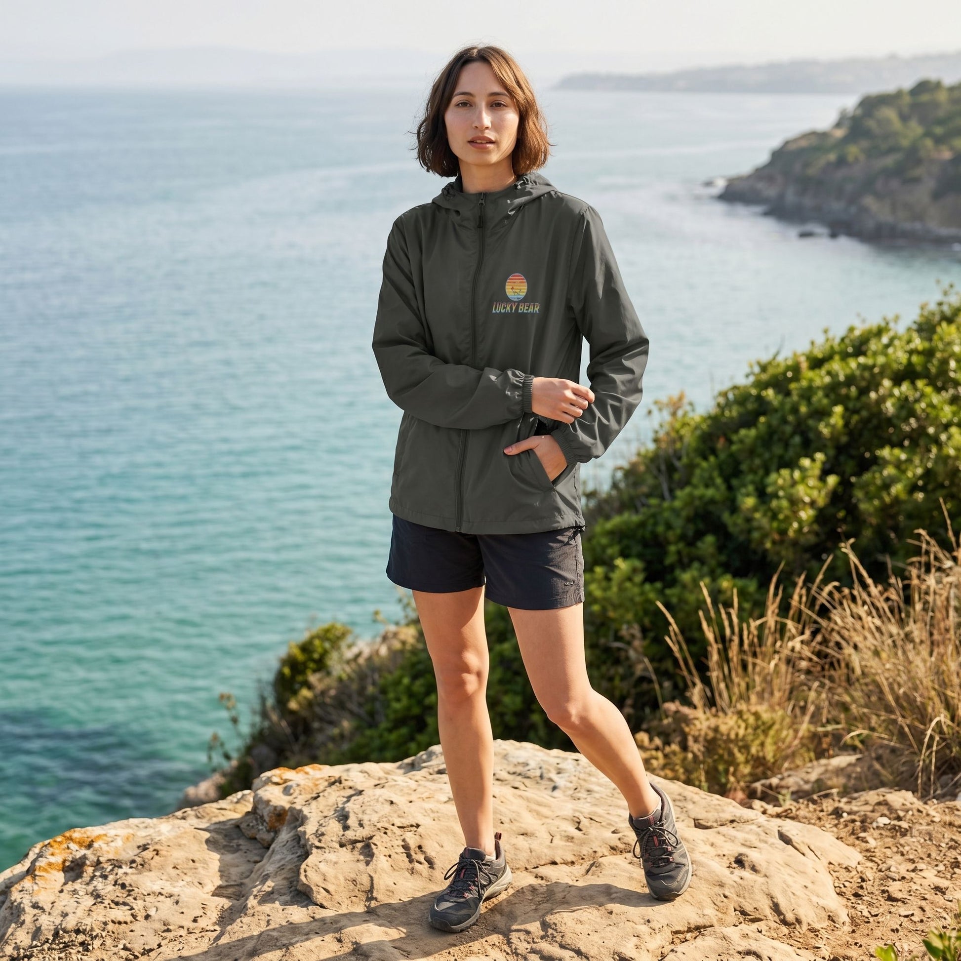 Full-body shot of a brunette female model outdoors wearing a lightweight, water-resistant windbreaker overlooking the ocean, highlighting its design and features.