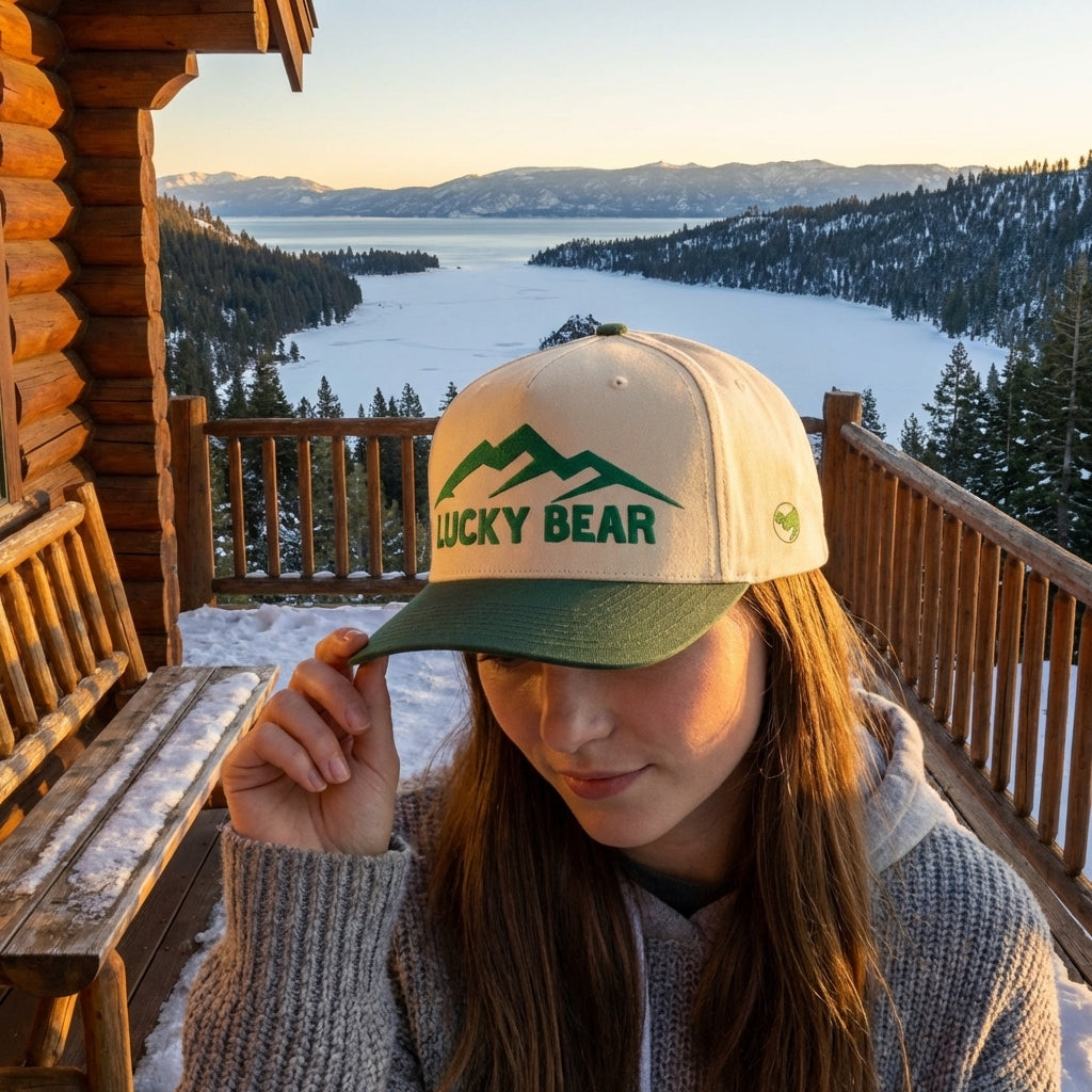 Model seated outdoors near mountain cabin wearing green Sierra 5 panel cap during golden hour.