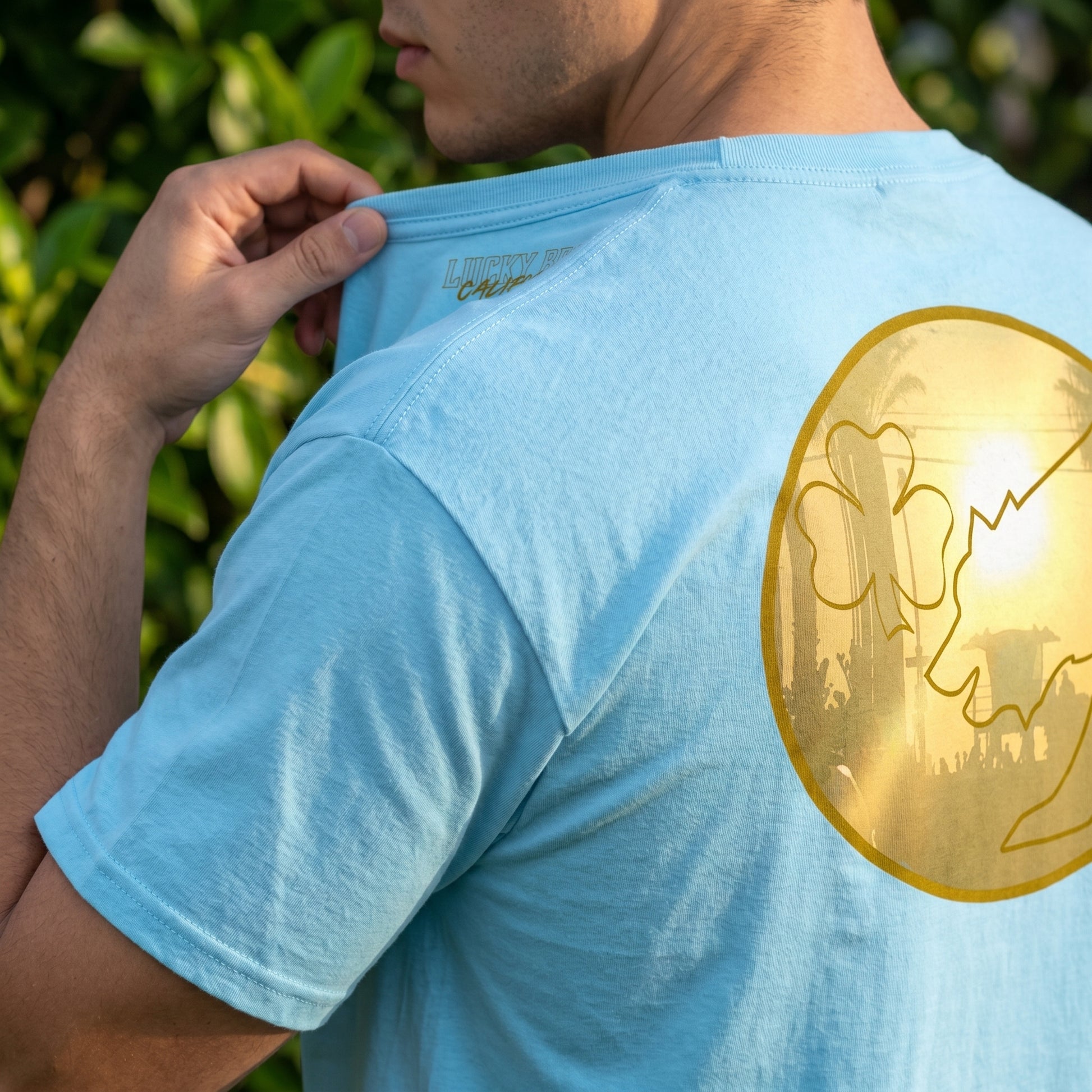 Macro view of San Diego Sunset men's cotton tee highlighting fabric weave and stitching, outdoors with greenery.