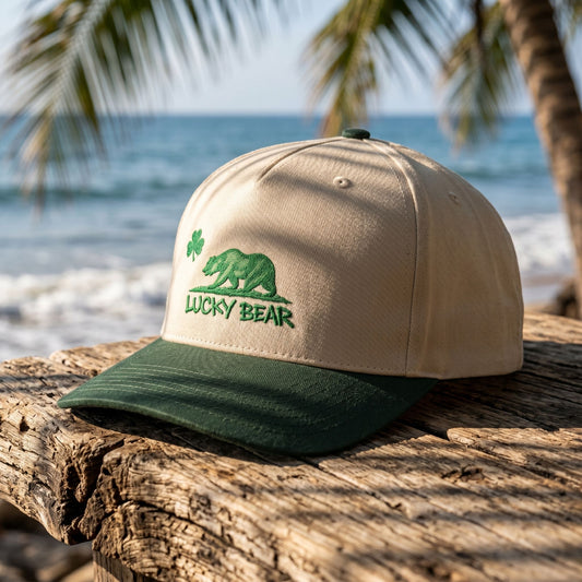 Close-up of Lucky Republic 5 panel cap on rustic wood near ocean, detailed spray-painted bear design framed by palm-shadowed sunlight.