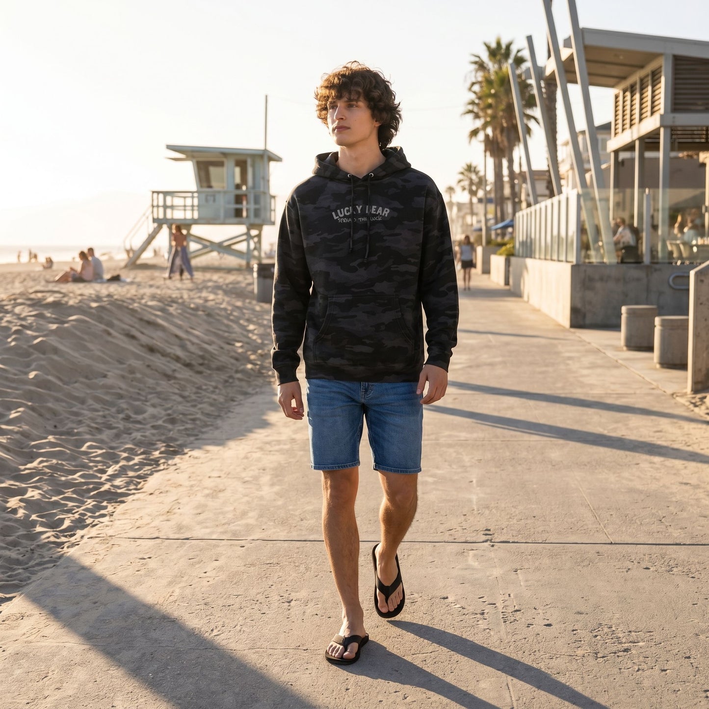 Model walking confidently on concrete beach sidewalk wearing a heavyweight hoodie, blue shorts, and flip flops, showcasing relaxed fit and durability.