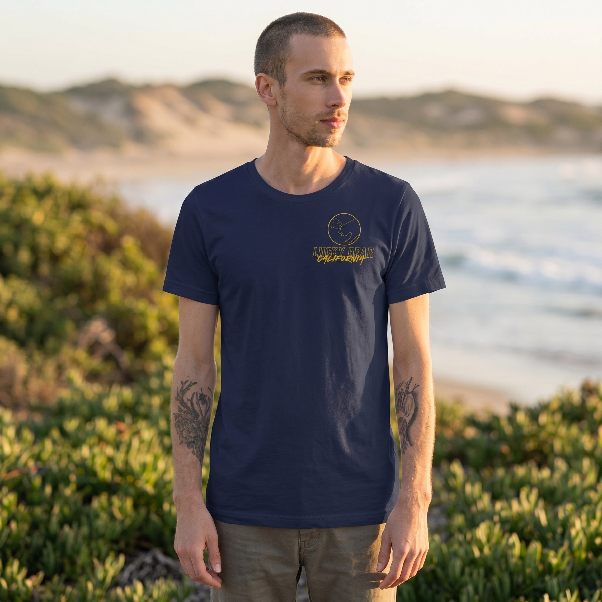 Full-body shot of a model in the Stay Golden Lucky Bear t-shirt on the beach, showing fit and variety of sizes with scenic natural background.