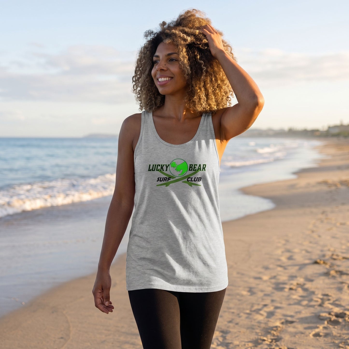 Full-body shot of a model wearing surf club logo tank top on a sandy beach during sunrise, with waves and sky in the background.