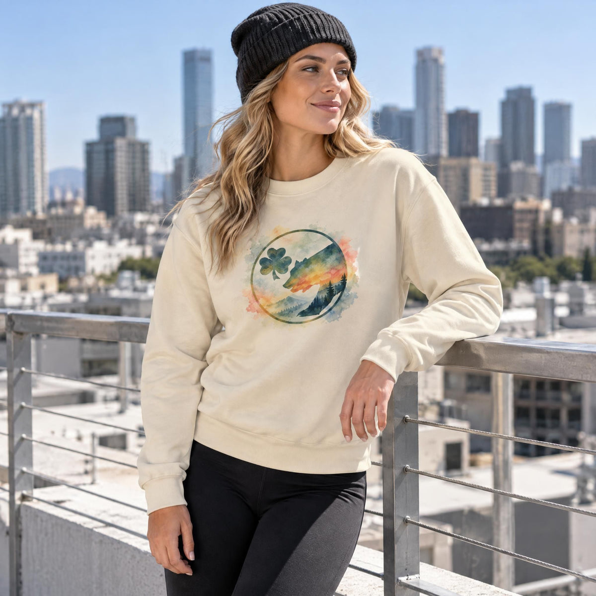Woman wearing a mountain sunset sweatshirt and beanie standing against a railing on an urban rooftop.