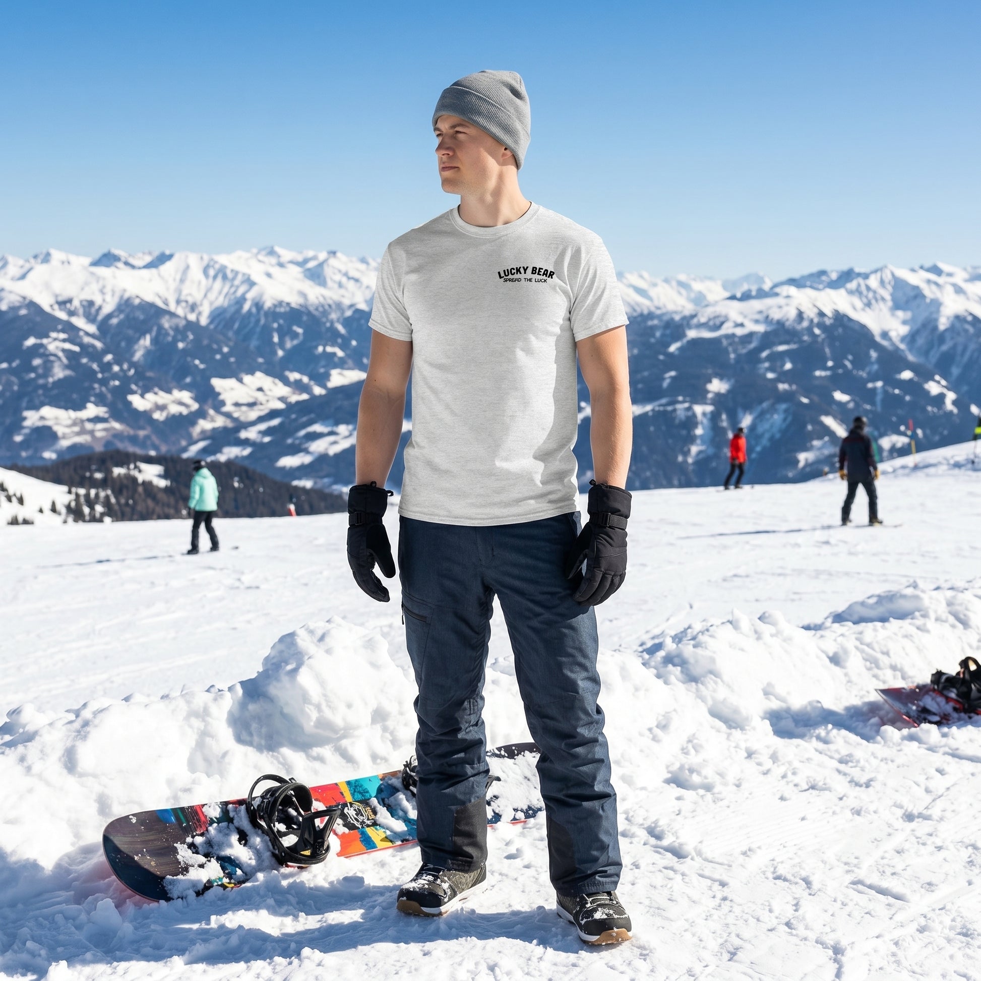 Male model wearing Lucky Bear snow day tee on snowy mountain with snowboard nearby in natural winter scene.