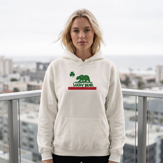 Blonde female model wearing a white Lucky Republic hoodie featuring a bear and shamrock graphic on a modern balcony.