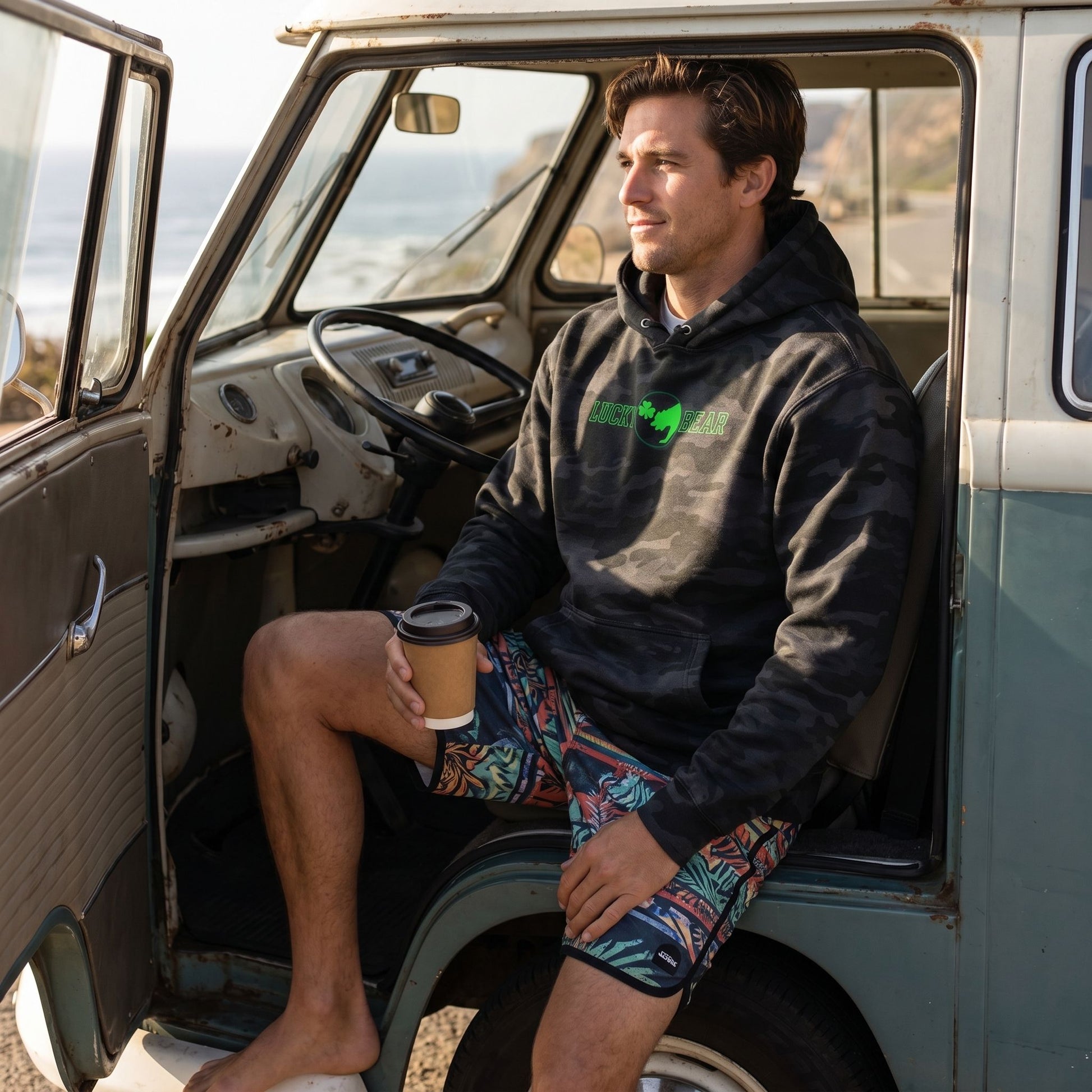 Male model in vintage Volkswagen van wearing camouflage Lucky Bear hoodie, relaxed fit, holding coffee, showcasing durability and comfort.