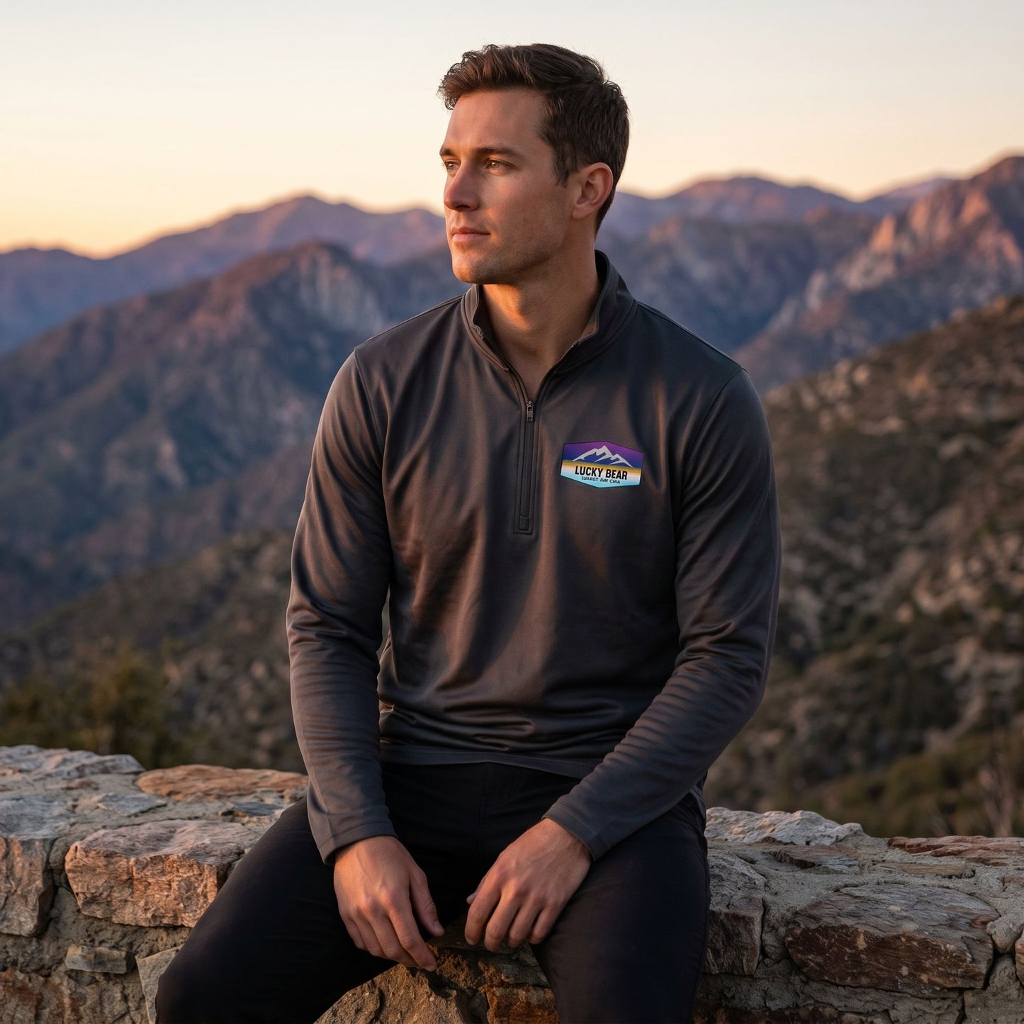 Model seated on stone wall at sunset wearing Sierra Sunset pullover with mountain range background at dusk.
