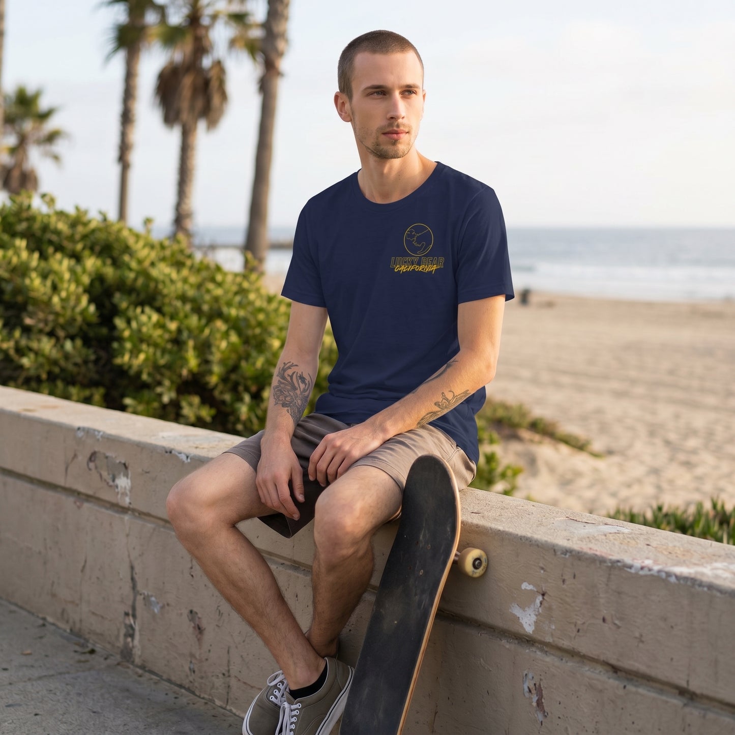Model sitting with skateboard on a concrete wall wearing the Stay Golden Lucky Bear t-shirt, displayed outdoors with natural scenery.