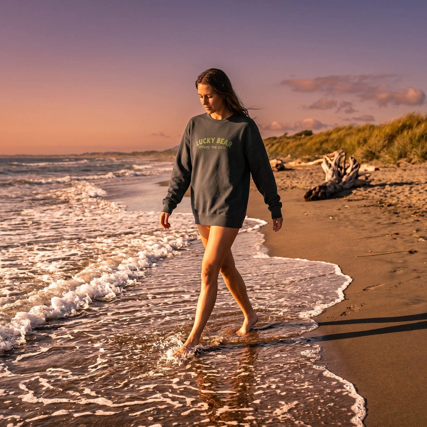 Model walking along shoreline during golden hour wearing Spread the Luck Sweatshirt, highlighting its relaxed fit and fabric texture.