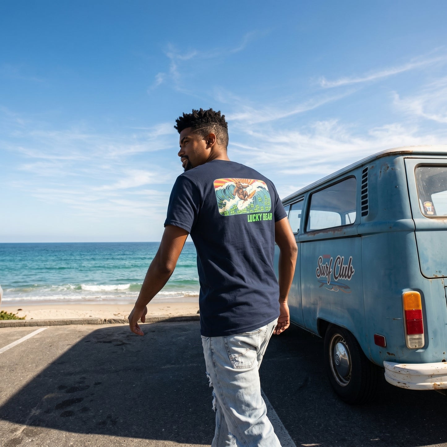 Model wearing Retro Surfing Bear Tee next to vintage VW bus at beachside parking lot with ocean horizon in background, capturing coastal lifestyle and vibrant colors.