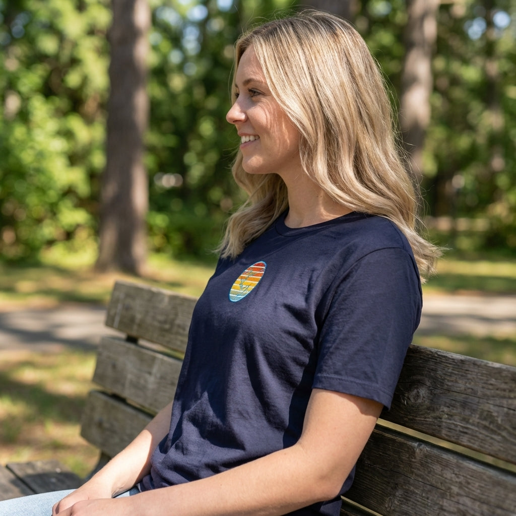 Side profile of woman sitting on bench in park wearing relaxed fit sunset T-Shirt, emphasizing fabric texture, logo, and casual style.