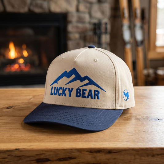 Blue Sierra 5 panel hat on wooden surface with rustic indoor background of vintage skis and lit fireplace, highlighting structured front panel and adjustable snap closure.