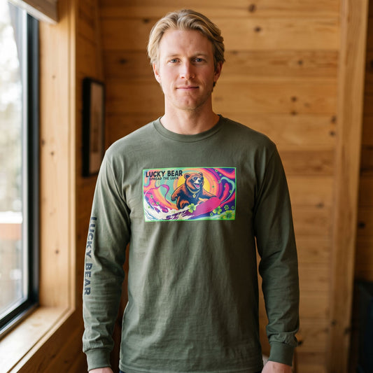 Blonde male model wearing a long-sleeve cotton shirt featuring a colorful surfing bear graphic print in a mountain cabin setting.