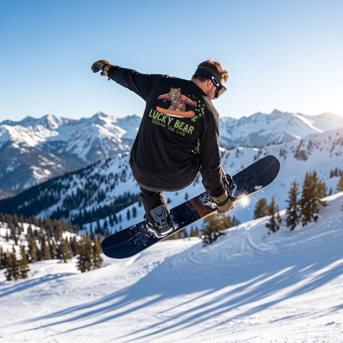 Male snowboarder in Sierra Shredding Bear long sleeve shirt mid-air against snowy mountain backdrop.