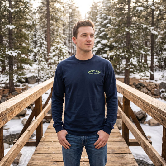 Male model wearing Sierra Shredding Bear long sleeve shirt on rustic wooden bridge in snow-dusted pine forest.