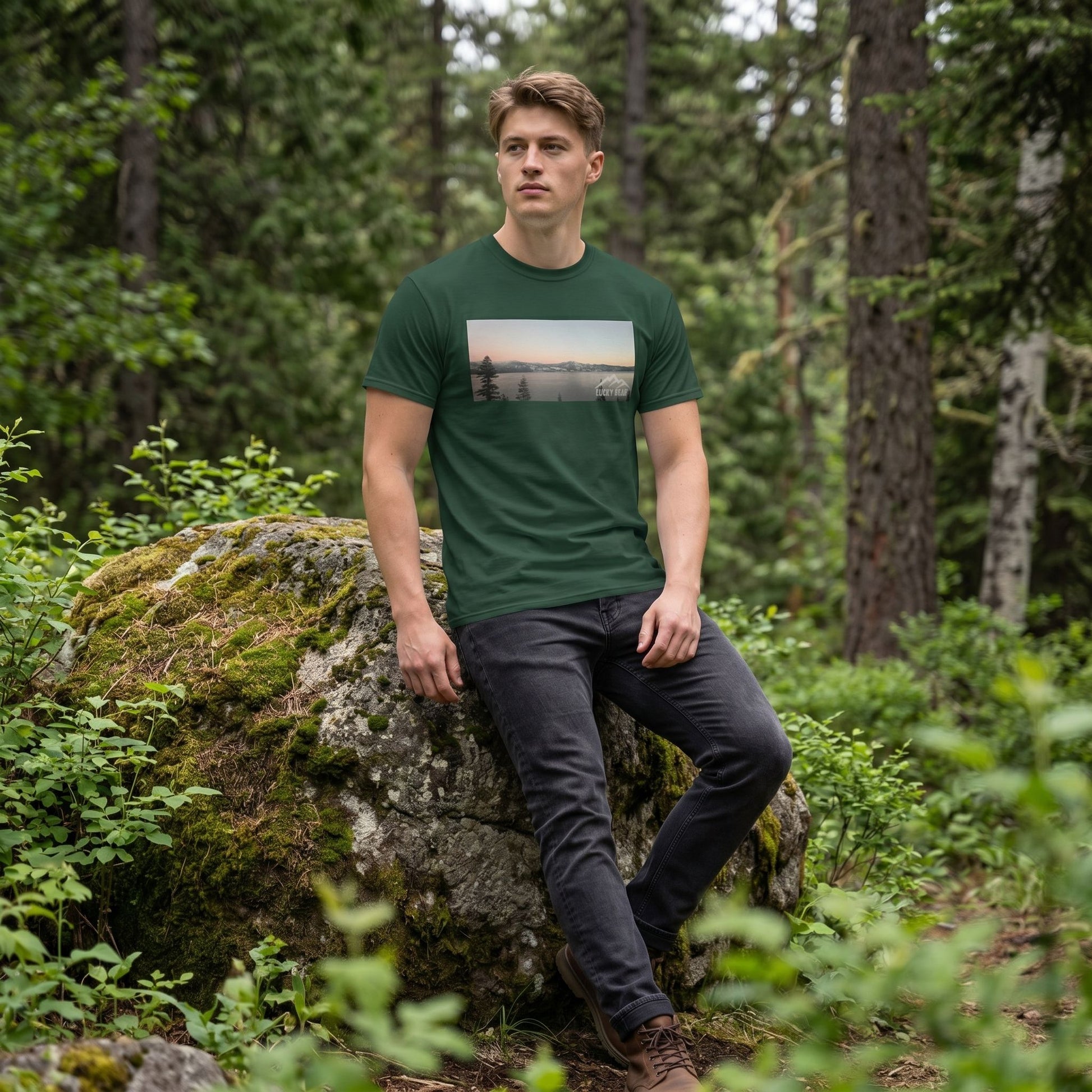 Full-body shot of model in forest green Sierra Sunset Tee leaning on mossy rock in lush forest clearing with soft diffuse lighting.