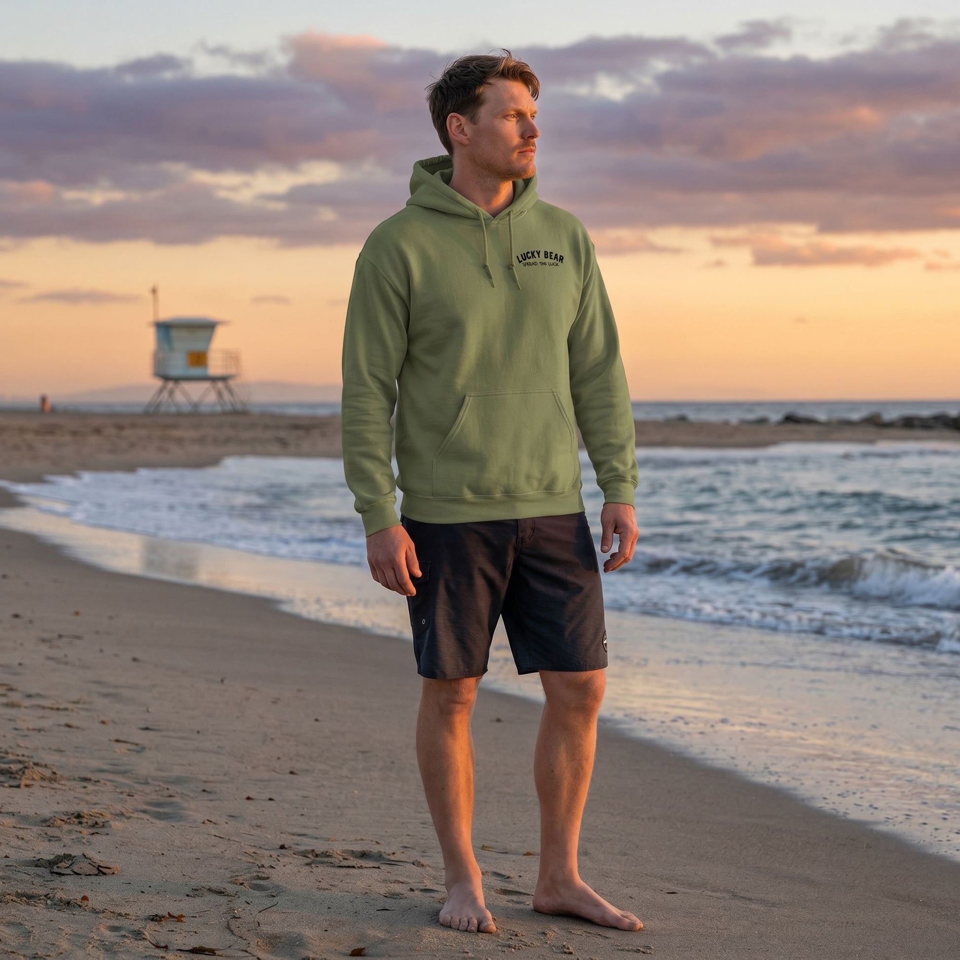 Full-body shot of the Spread the Luck Stealth Hoodie styled with boardshorts outdoors on a beach during sunset, highlighting fabric texture and fit.