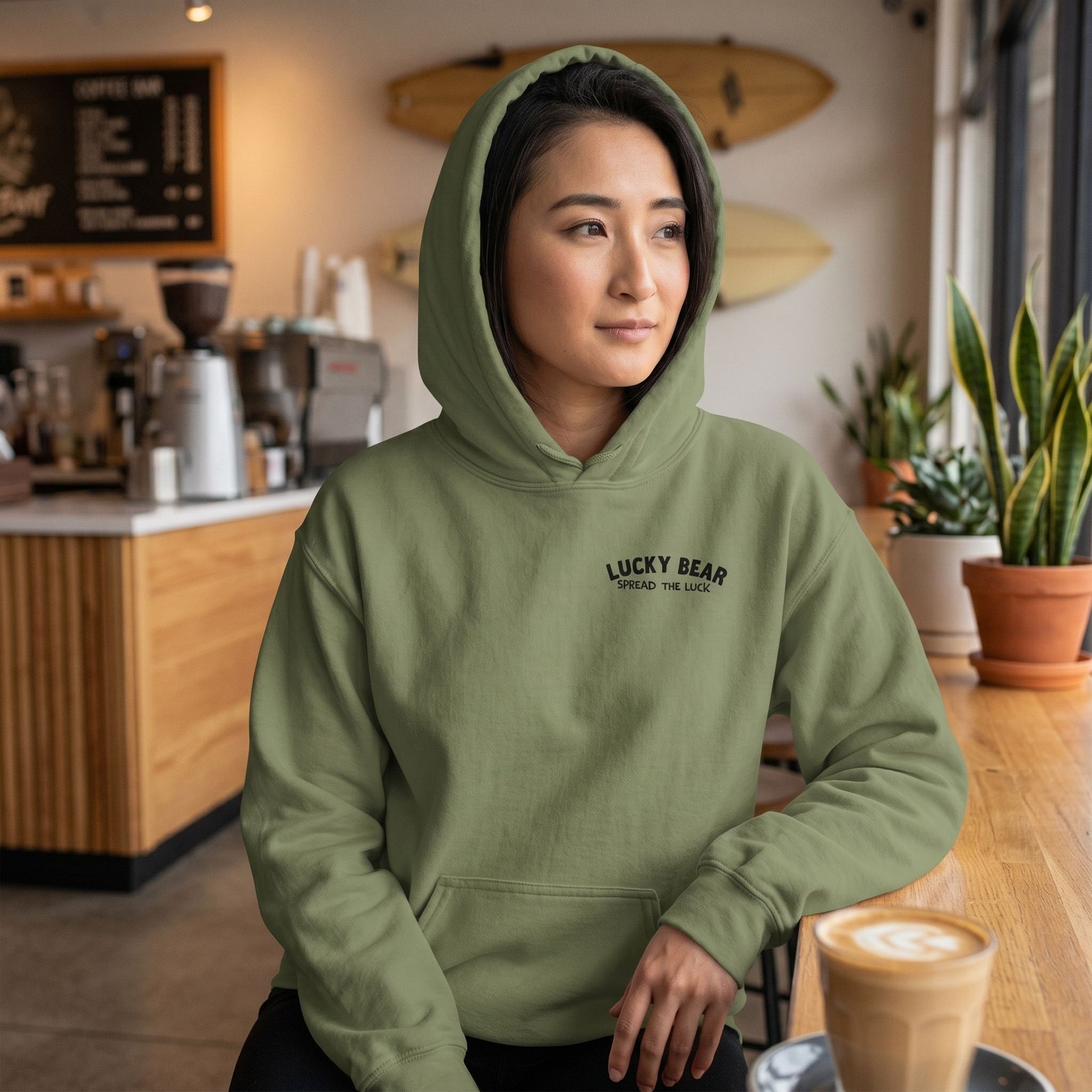 Relaxed model wearing Spread the Luck Stealth Hoodie in a warmly lit modern surf-themed coffee shop, emphasizing the soft texture and fit.