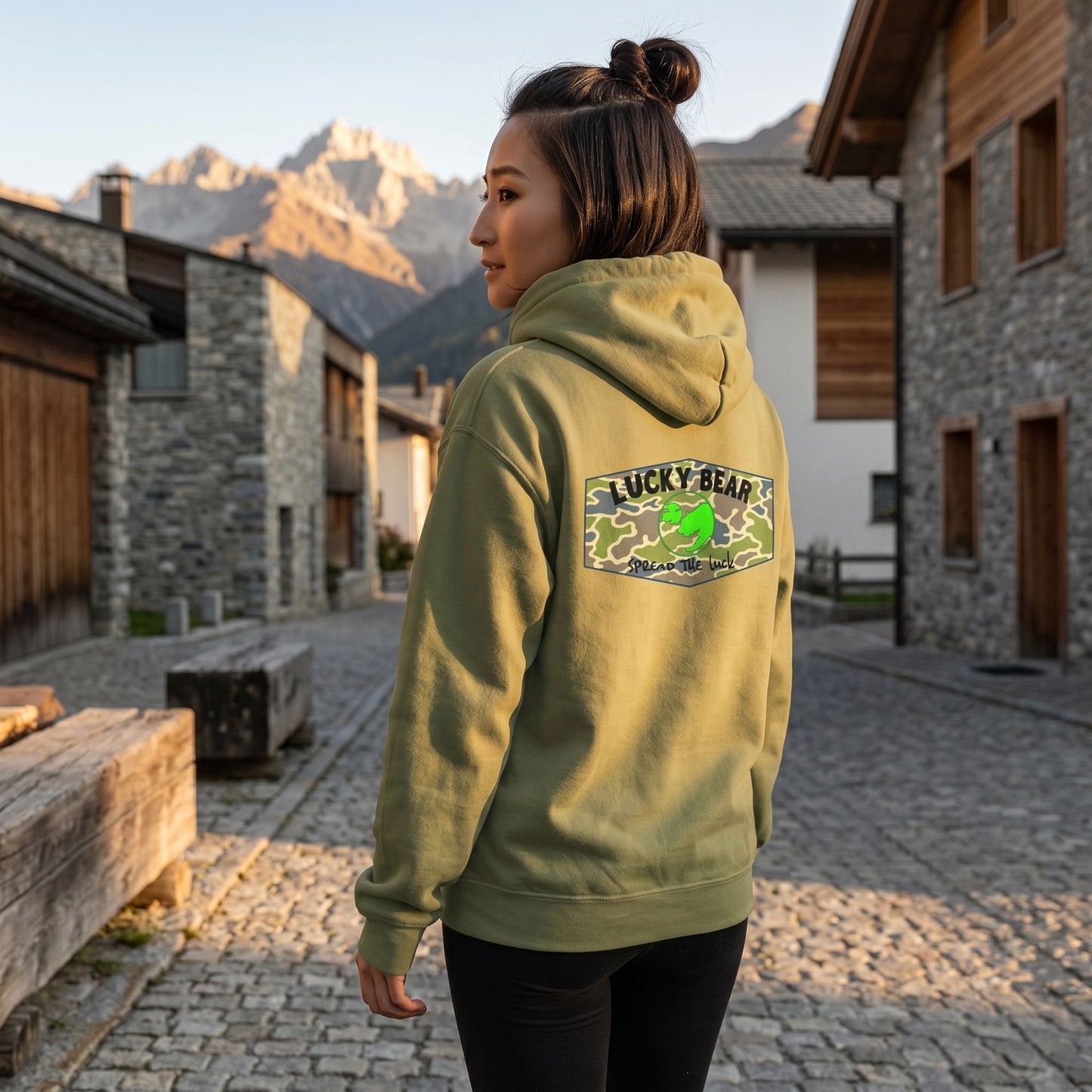 Model wearing Spread the Luck Stealth Hoodie walking in a mountain village during golden hour, showcasing fabric smoothness and trendy style.