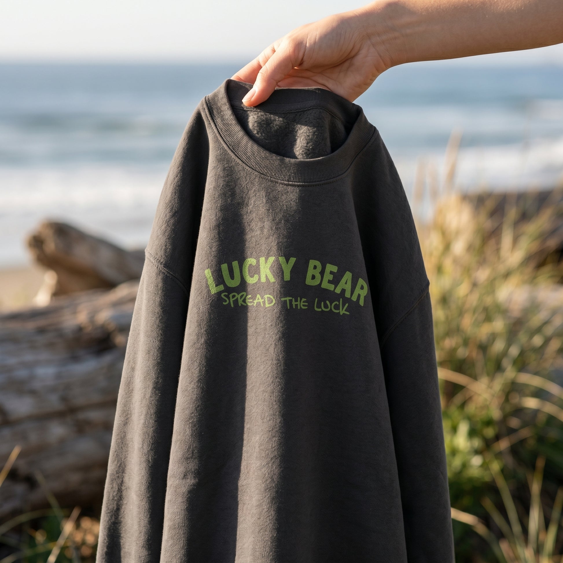 Close-up of the Spread the Luck Sweatshirt hanging from a model's hand, showcasing fabric texture and printed logo against a blurred oceanfront background in warm natural sunlight.