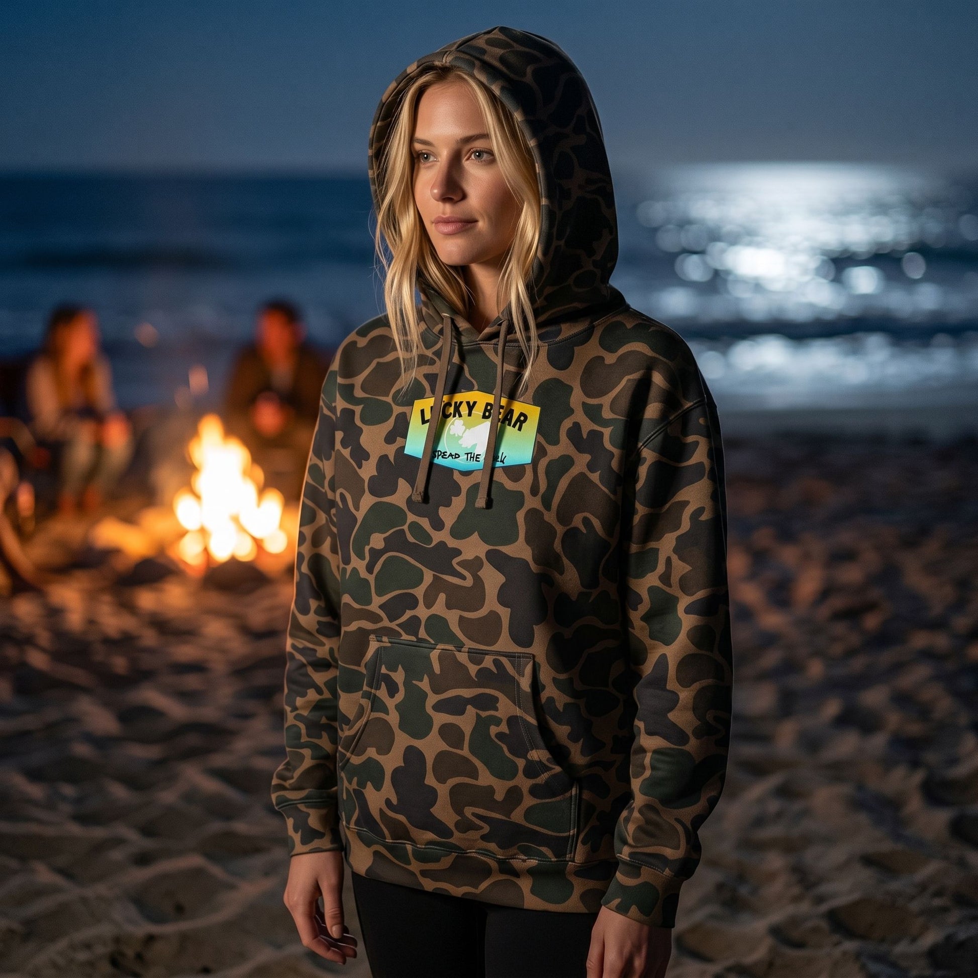 Female model wearing camouflage Summer Sunset Hoodie with hood up on beach after dark, highlighting textured fabric and high-quality stitching near bonfire and moonlit ocean.
