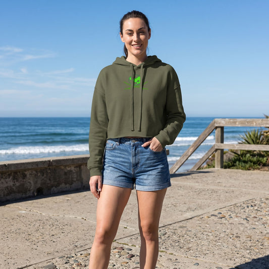 Full-body of a model wearing a green Surf Club Logo Crop Hoodie and jean shorts on a textured concrete patio with ocean waves in the background and natural sunlight.