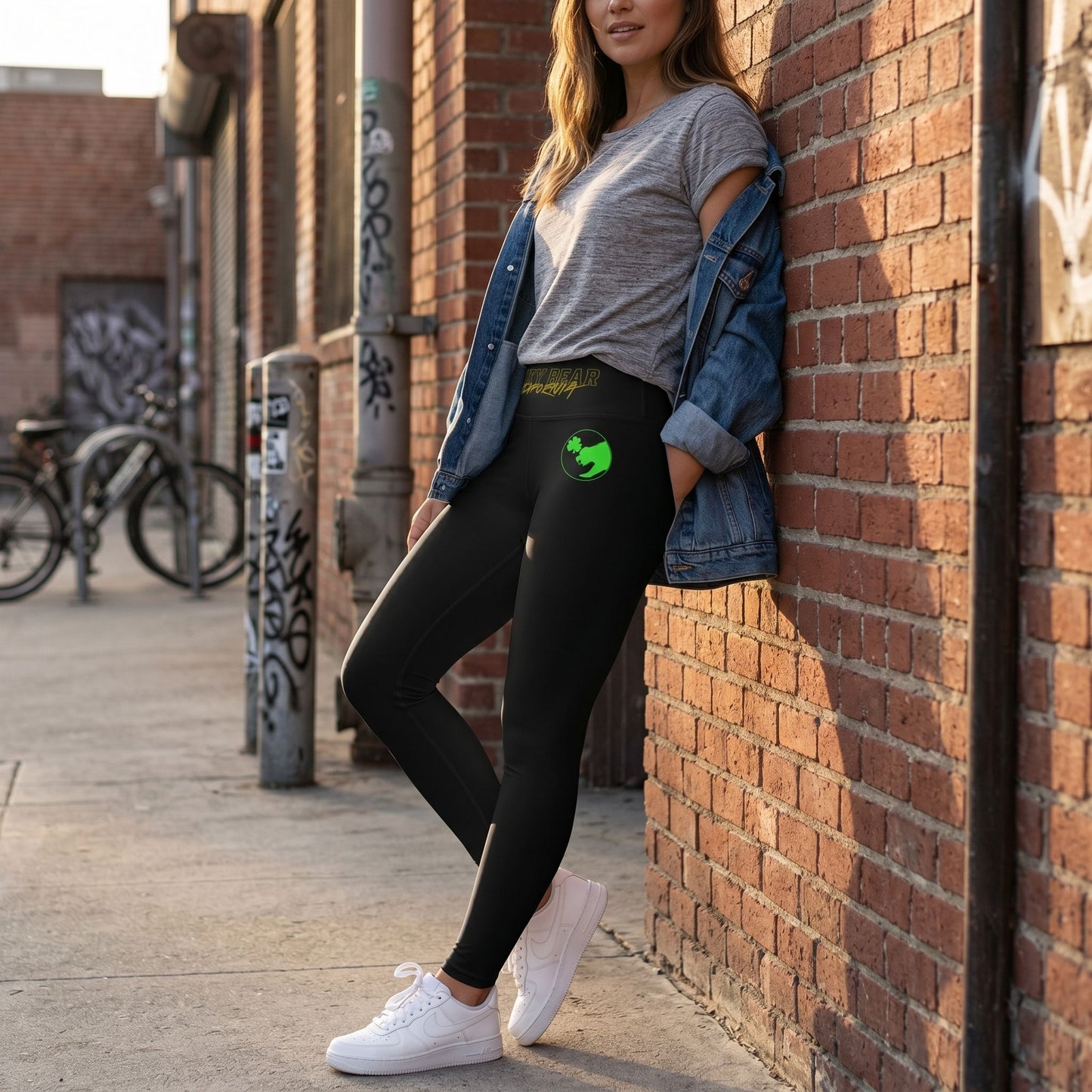 Waist-up of a model in black yoga leggings with Shamrock Bear logo, leaning against a red brick wall during golden hour, showcasing a relaxed urban streetwear look.